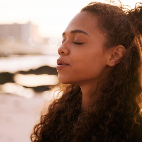 A person with long, curly hair stands on a beach at sunset, eyes closed and lips slightly parted, conveying a sense of peace and serenity.
